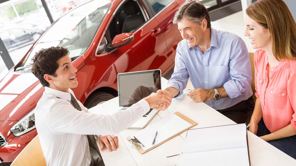 Ein Autoverkäufer schüttelt den Kunden die Hand in einem Autohaus in Coesfeld, im Hintergrund ist ein rotes Auto zu sehen und auf dem Tisch liegen Unterlagen.